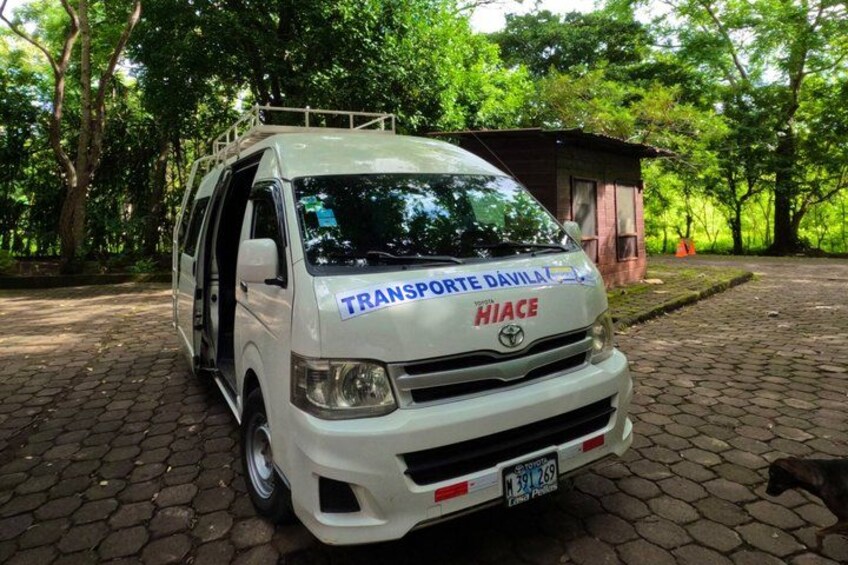 A white van with 'TRANSPORTE DÁVILA HIACE' on the front, parked on a cobblestone path surrounded by trees. 3