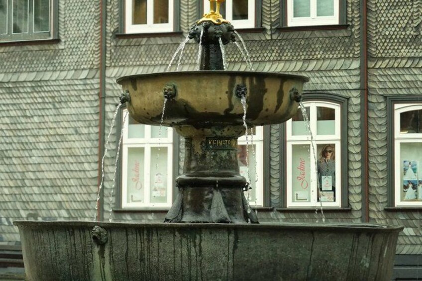 A historic fountain with water spouting from its top, set against a building with ornate window frames and a textured facade. 5
