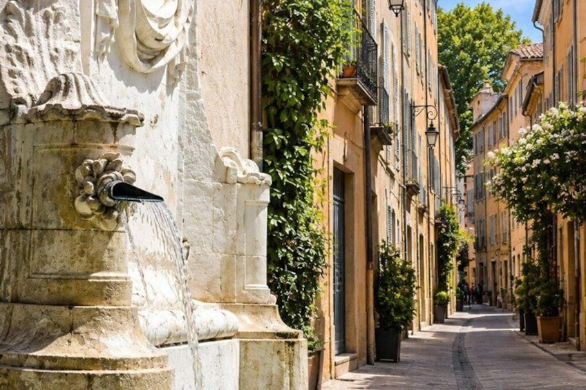 A stone fountain with water spouting from it, a narrow street lined with buildings, and a clear blue sky. 2