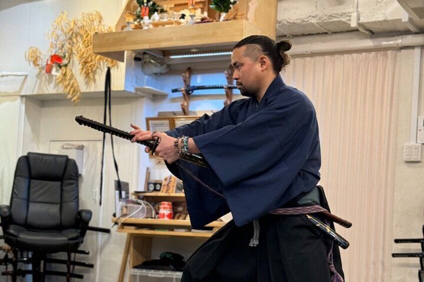 A person in traditional attire holding a sword, seated in a room with a desk and chair. 3