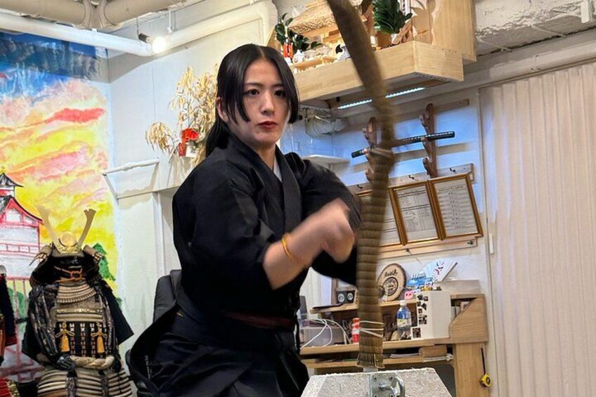 A person in a black outfit practicing martial arts with a wooden staff in a room with various decorations and a painting in the background. 1