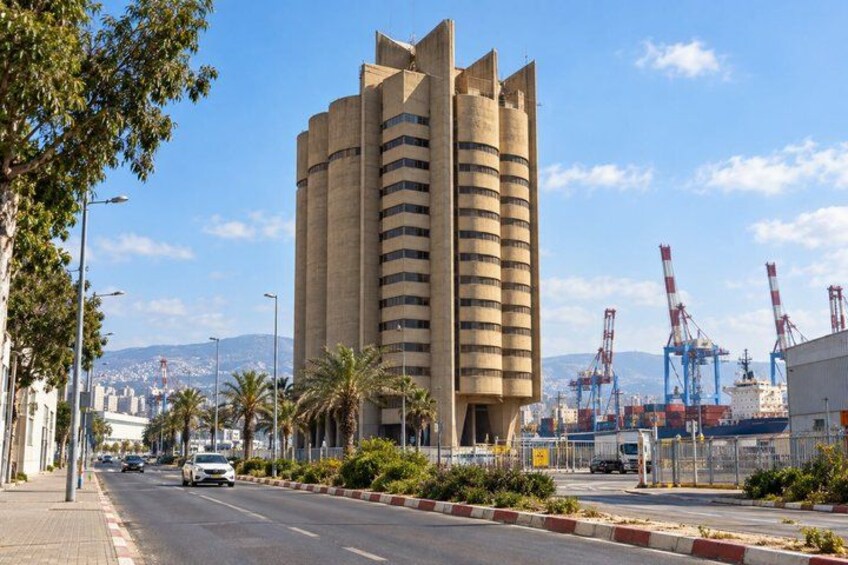 A tall building with a cylindrical shape, a road with palm trees, and a port with cranes in the background. 1