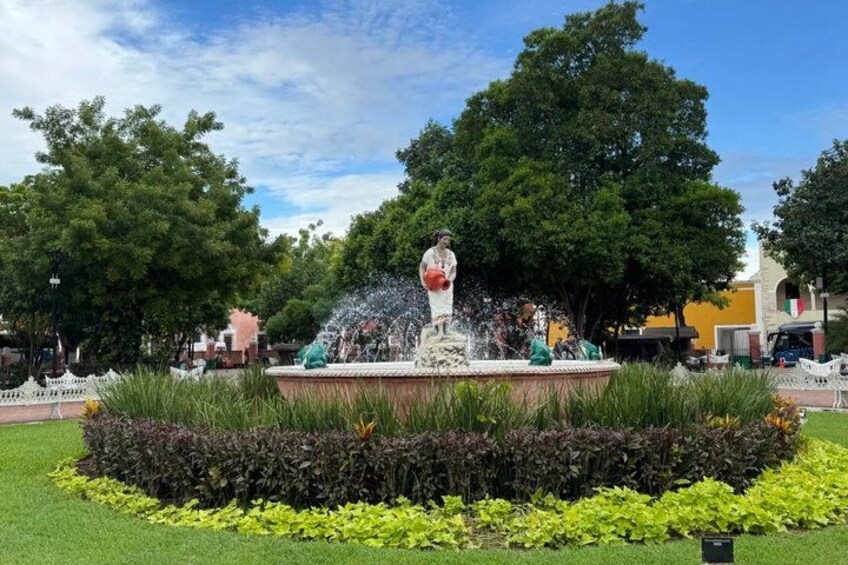 A fountain with a statue, surrounded by well-maintained grass and plants, with trees and buildings in the background. 5