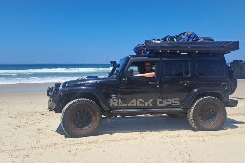 A black off-road vehicle with luggage on the roof parked on a sandy beach. 1