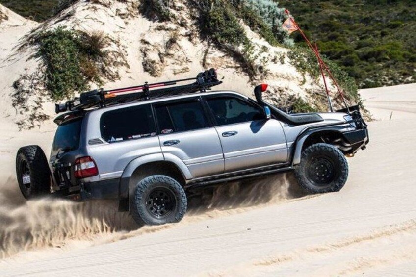 A silver SUV with off-road tires and a roof rack, driving on a sandy terrain with a rocky hill in the background. 4