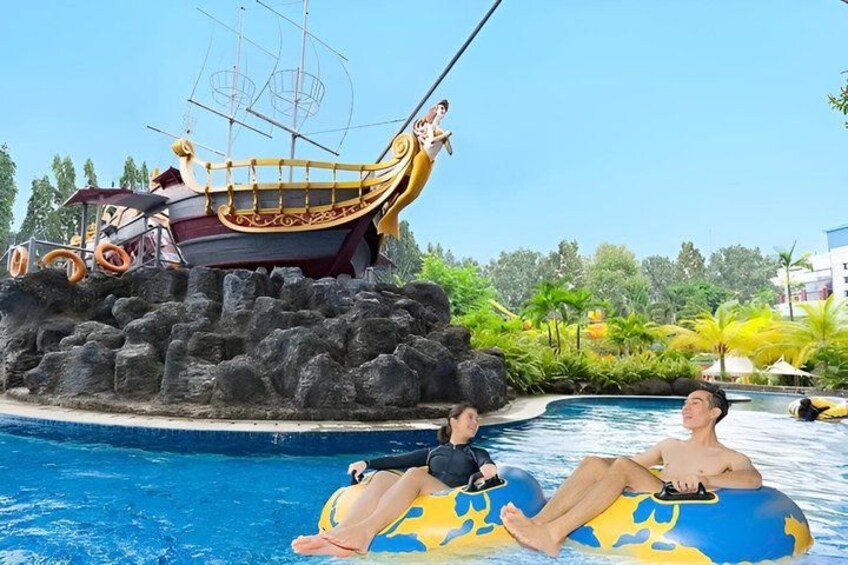A swimming pool with two people on inflatable tubes, a shipwreck, and tropical landscaping. 5