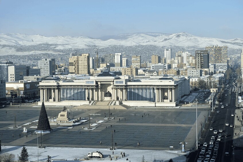 Un paisaje urbano con un edificio grande con columnas en primer plano, una cordillera nevada al fondo y una calle concurrida con coches. 2