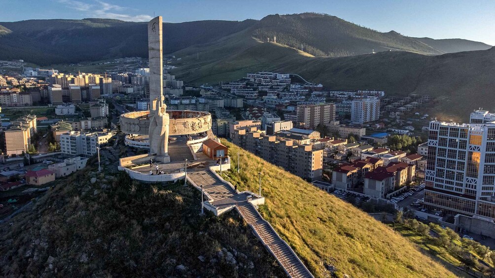 Un paisaje urbano con un monumento destacado en una colina, rodeado de edificios residenciales y un telón de fondo montañoso. 4