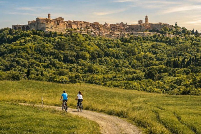 A rural landscape with a dirt path, two cyclists, and a historic town on a hill in the background. 1