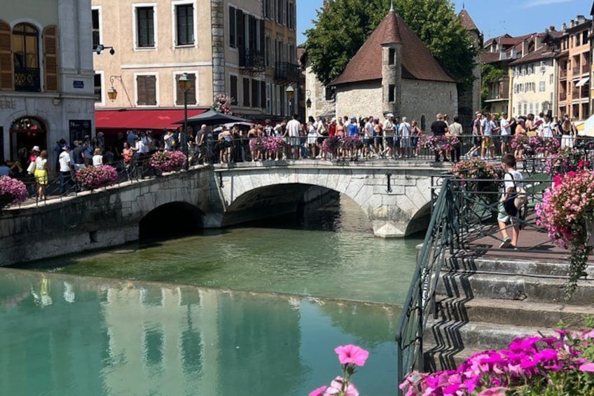 A stone bridge over a canal with people walking on it, surrounded by buildings and flowers. 1