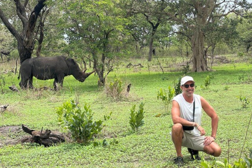 A person crouching in a grassy field with a rhinoceros in the background. 1