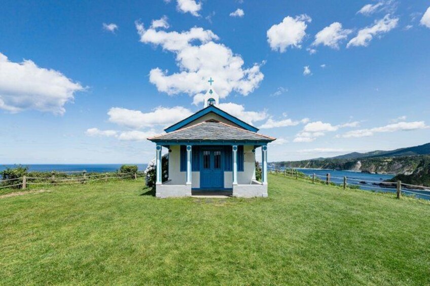 A small blue and white chapel with a cross on top, surrounded by a grassy field and a coastline. 3