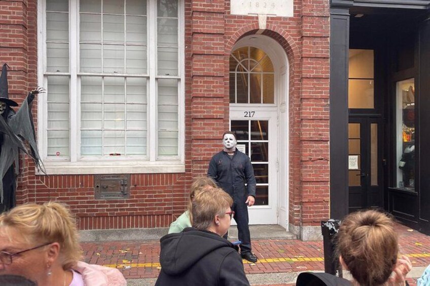 A person in a black coat and white mask standing in front of a brick building with a white door marked '217'. 1
