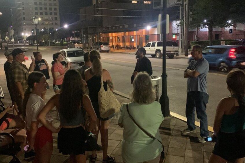 A group of people standing on a sidewalk at night, a street with cars, and buildings in the background. 3