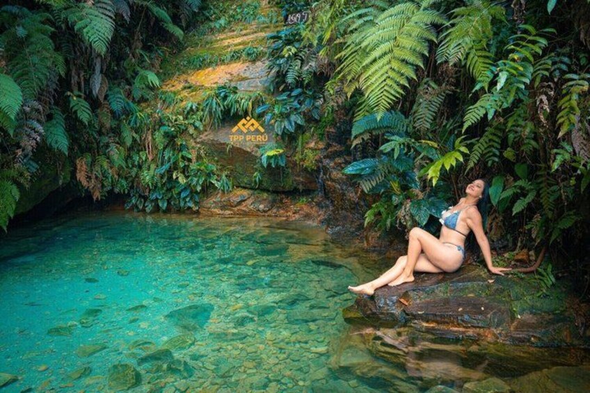 A person sitting on a rock by a clear water body surrounded by dense green foliage. 1
