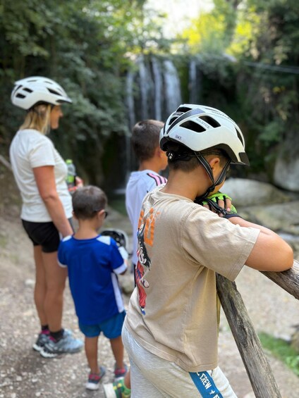A person wearing a helmet and a beige t-shirt with a graphic design, standing on a path with a waterfall in the background. 3