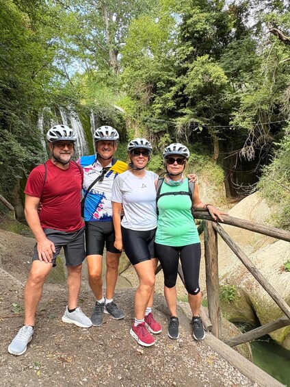 Four people wearing helmets and cycling attire standing on a bridge with greenery in the background. 5