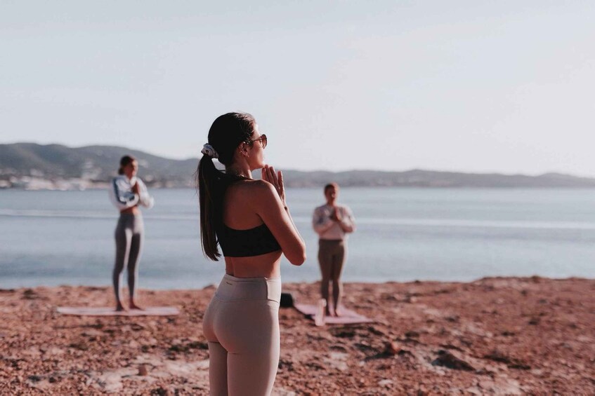 Tre persone praticano yoga su una spiaggia con un mare calmo e colline sullo sfondo. 3