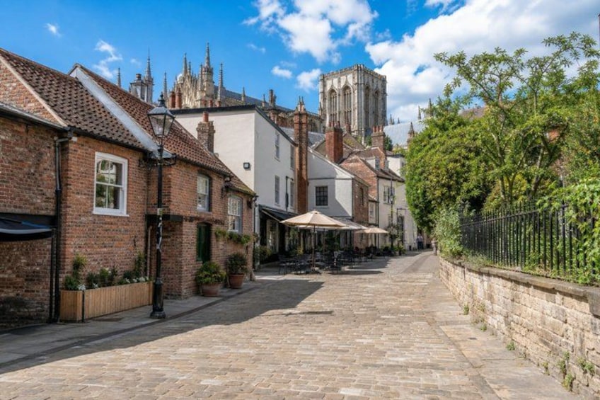 A cobblestone street lined with brick houses, a church tower, and outdoor seating at a cafe. 2