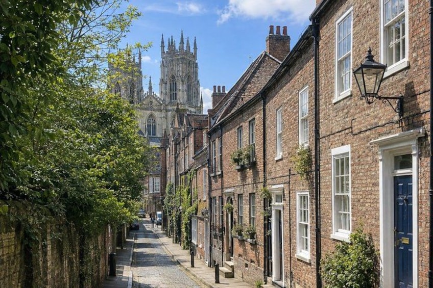 A narrow street lined with brick buildings, a church spire in the distance, and a tree on the left. 3