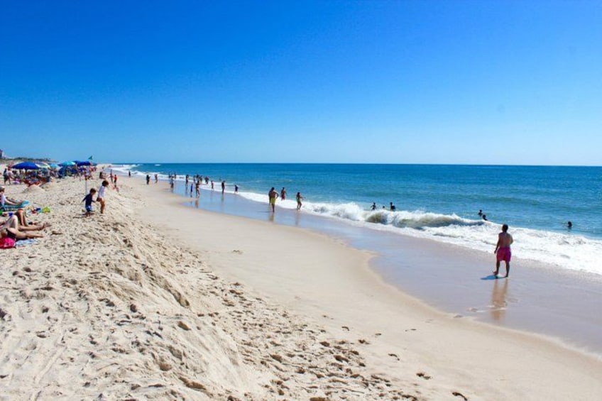 A sandy beach with people walking and standing, and umbrellas in the background. 2