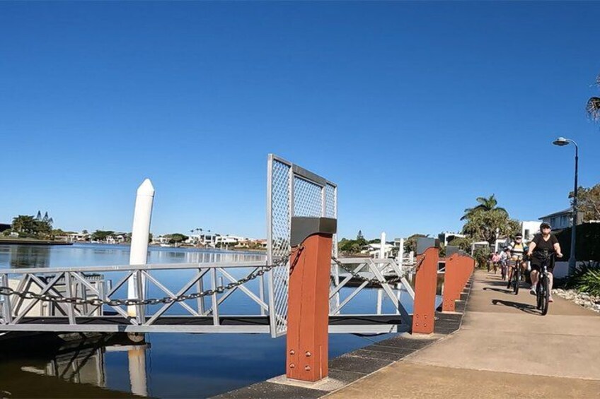 A pedestrian bridge over water with metal railings and red pillars, a clear blue sky, and a cyclist in the distance. 2