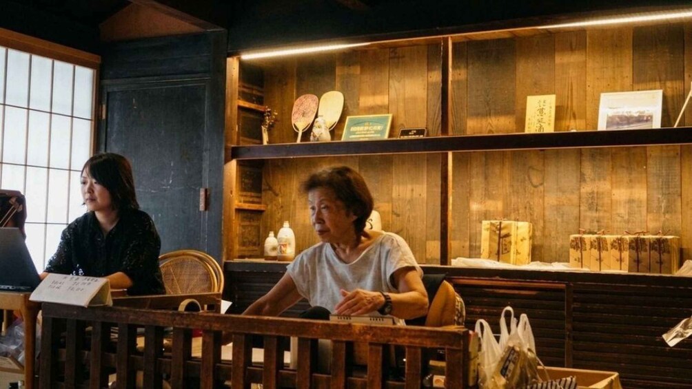 Two people seated at a wooden table in a room with shelves containing various items. 3