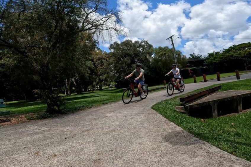 Two cyclists on a paved path in a park with trees and grass. 2