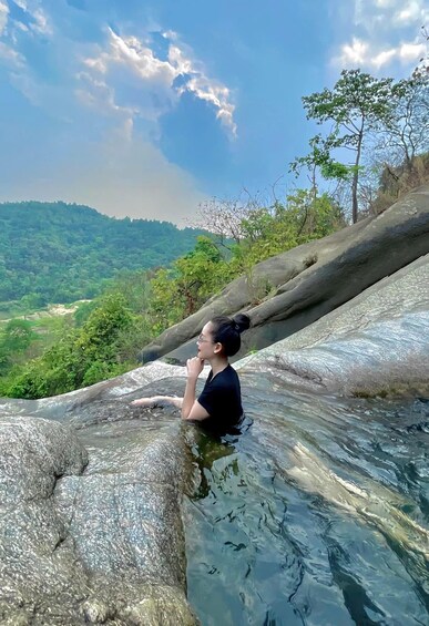 A person sitting on a rock by a clear stream, surrounded by greenery and a mountainous landscape. 4