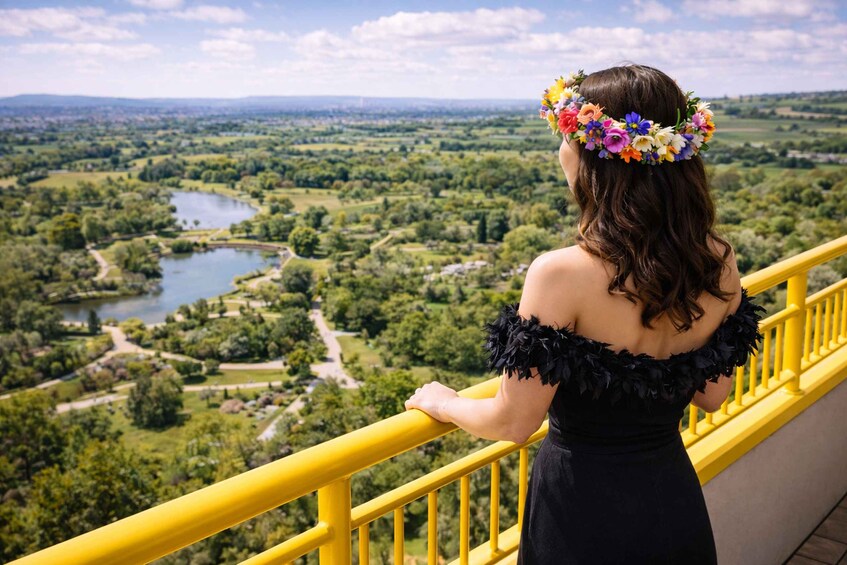 Una persona con vestido negro y una corona de flores, de pie sobre una barandilla amarilla que da vista a un paisaje verde con un río. 3