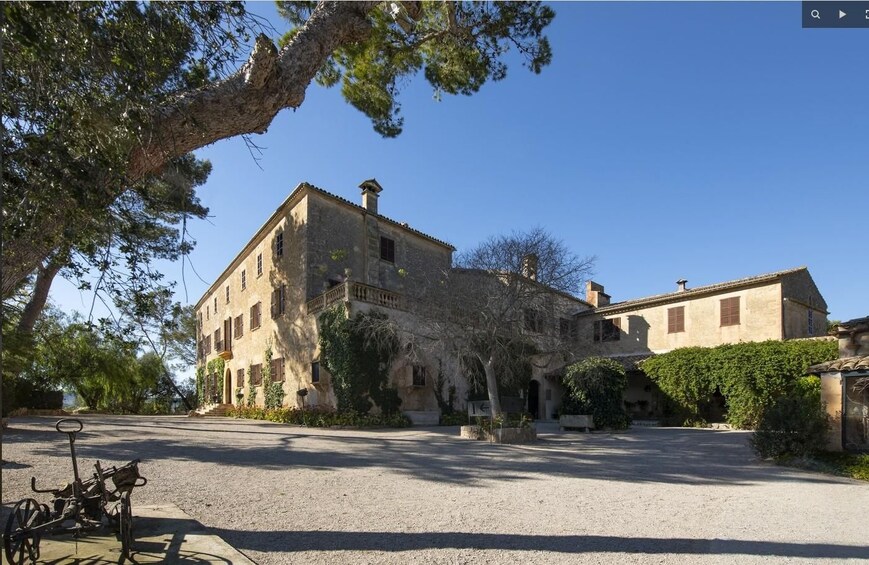 A historic stone building with a courtyard, surrounded by trees and a gravel path. 1