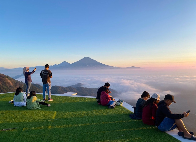 A group of people sitting and standing on a grassy hill overlooking a mountain and clouds. 2