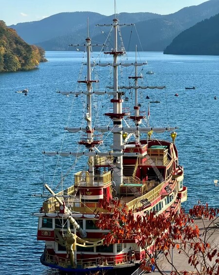 A large, multi-decked riverboat with multiple masts and sails, docked near a shoreline with autumn-colored trees and hills in the background. 1