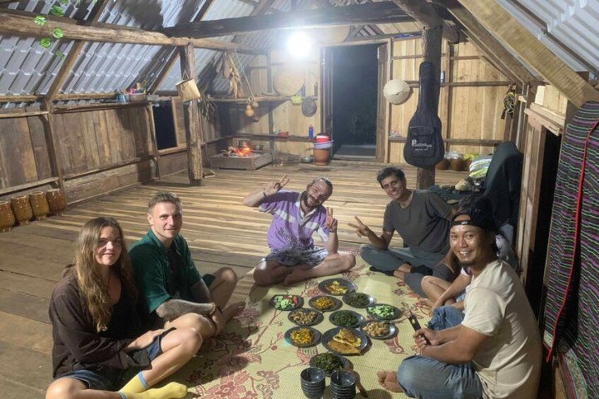 A group of people sitting on a wooden floor inside a rustic wooden structure with a pitched roof, surrounded by various dishes and a guitar hanging on the wall. 5