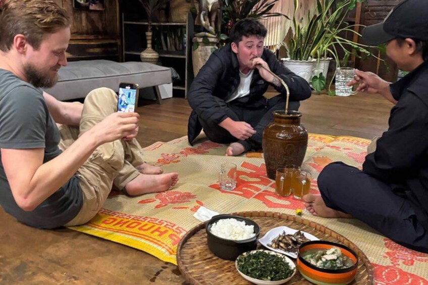 Three people sitting on a rug, one holding a smartphone, another drinking from a jar, and a third holding a glass. There are bowls of food and a large jar on the table. 1