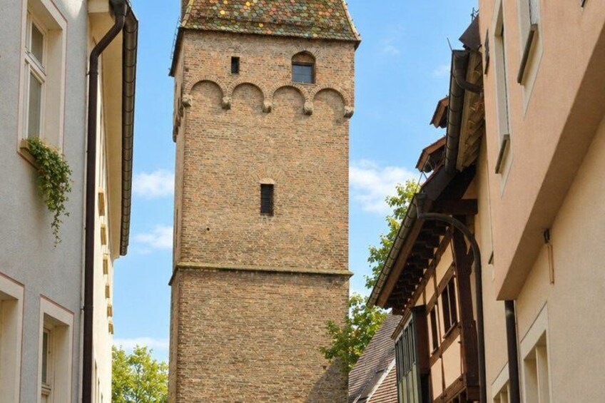 A tall brick tower with arched windows and a small balcony, surrounded by buildings with traditional architecture. 4