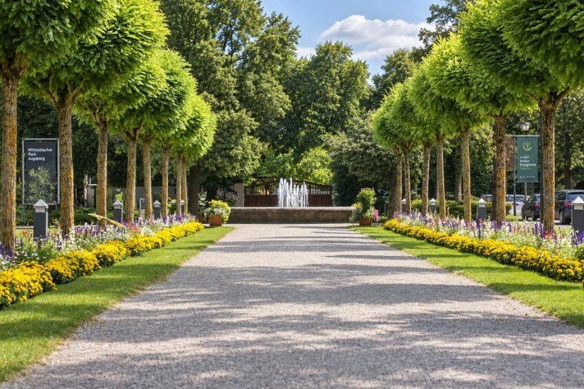 A pathway lined with trees and flowers leading to a fountain. 4
