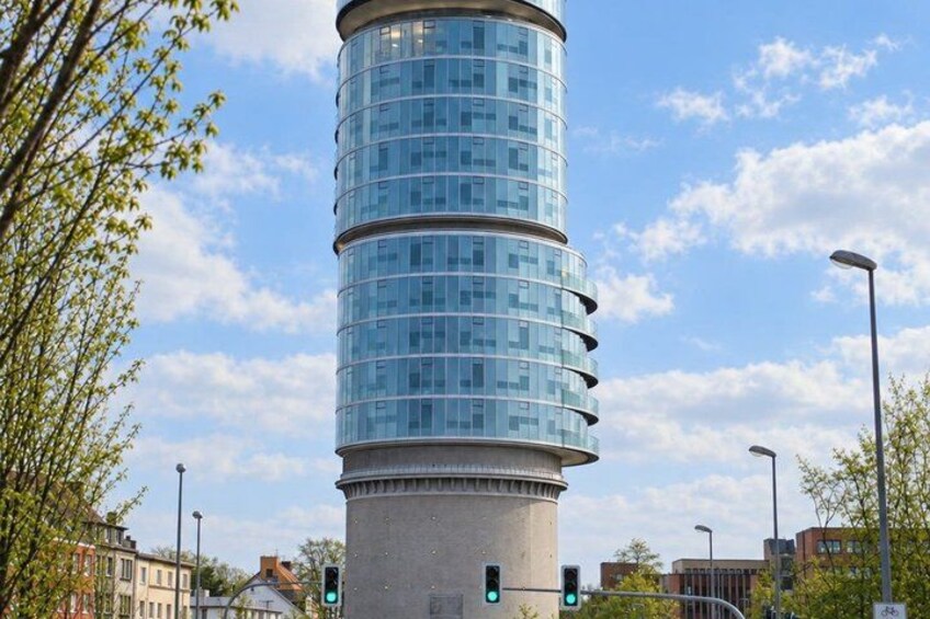 A cylindrical glass tower with multiple floors, surrounded by trees and urban buildings. 3