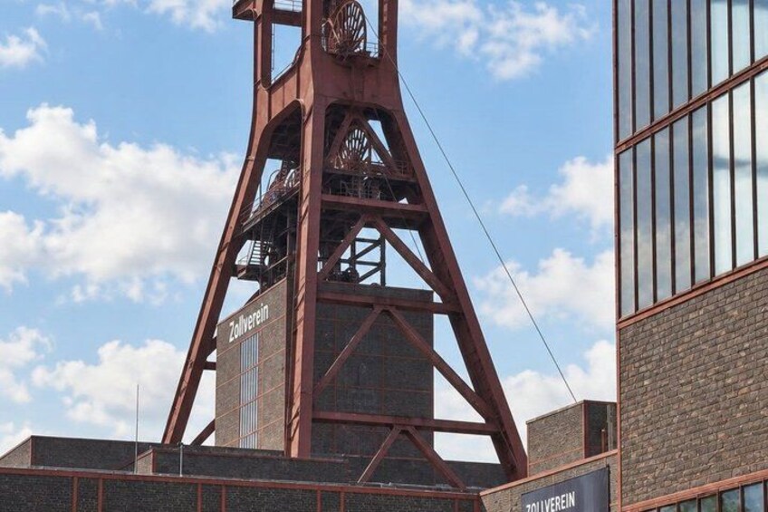 A tall, rust-colored metal structure with the name 'Zollverein' on its side, set against a partly cloudy sky. 5