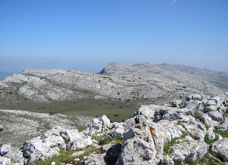 A rocky mountain landscape with sparse vegetation and distant peaks under a clear sky. 3