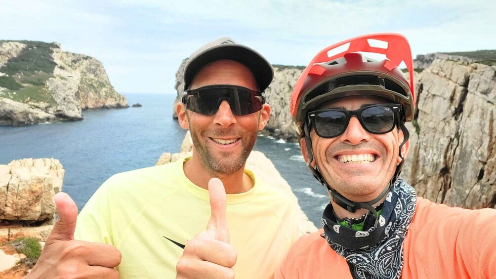 Two people giving thumbs up, one in a yellow shirt and the other in a red helmet, with a rocky coastline and sea in the background. 4