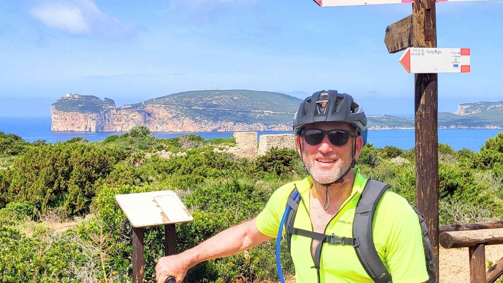 A person wearing a helmet and a neon vest, standing in front of a signpost with a coastal landscape in the background. 1