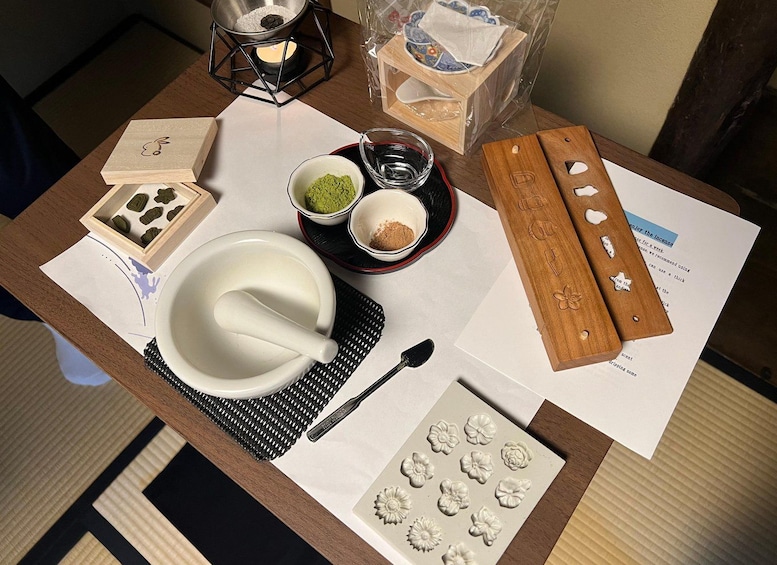 A table with a wooden cutting board, a bowl of green paste, a mortar and pestle, a spoon, a paper with flower-shaped molds, and a candle. 2