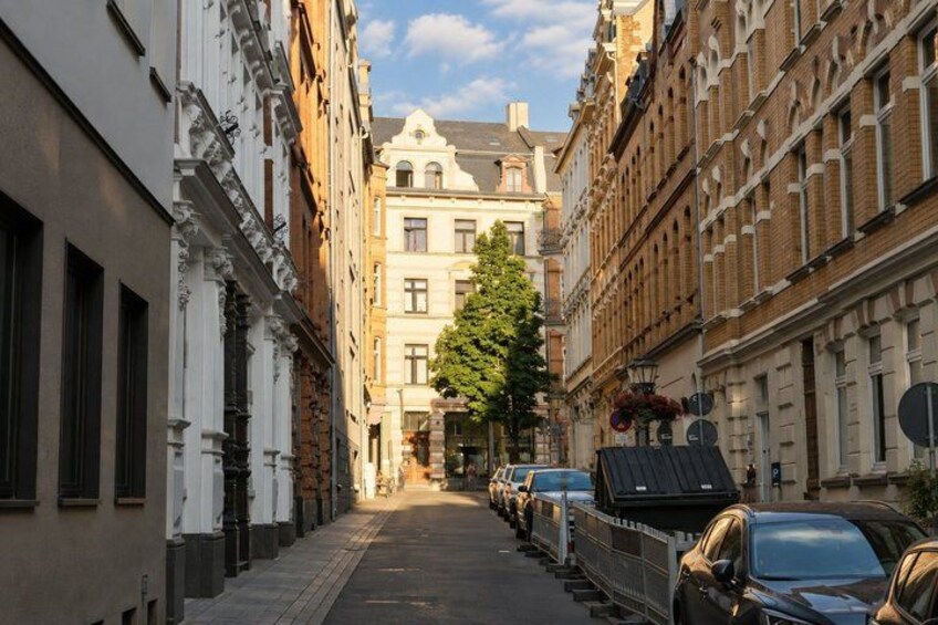 A narrow street lined with historic buildings, parked cars, and a tree in the center. 2