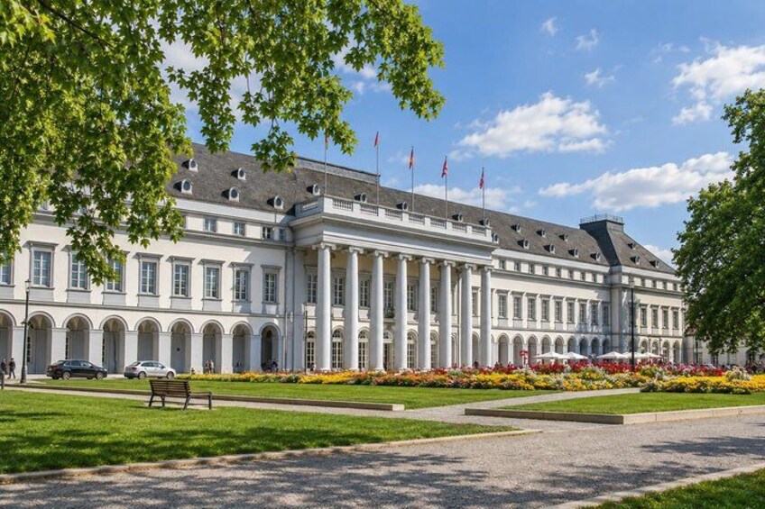 A large, classical building with columns, a paved courtyard, and a well-maintained garden with colorful flowers. 5