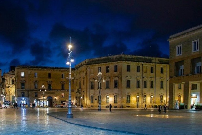 A nighttime urban square with historic buildings, street lamps, and a wet ground reflecting lights. 2