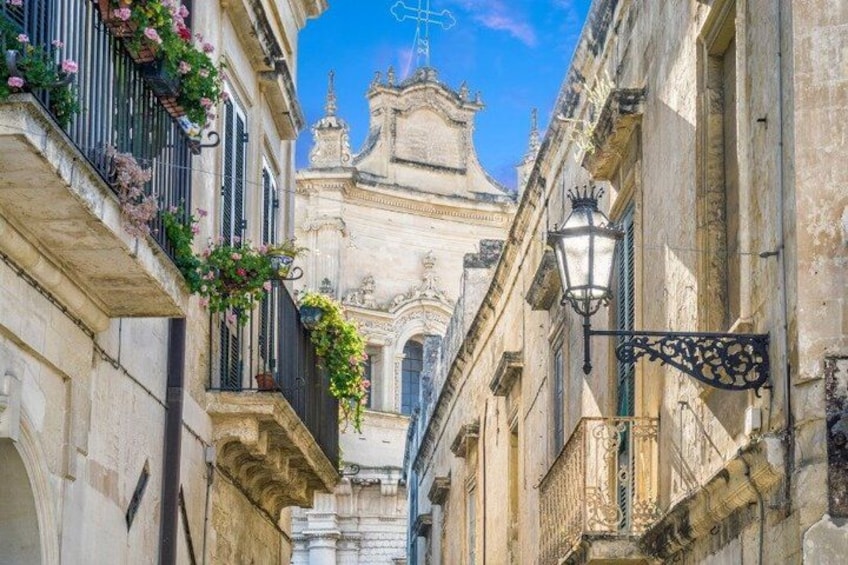 A narrow street with ornate buildings, a church with a bell tower, and hanging flower baskets. 5