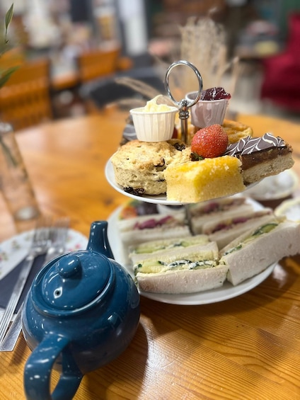 A tiered dessert tray with various cakes and pastries, a teapot, and a glass of water on a wooden table. 5