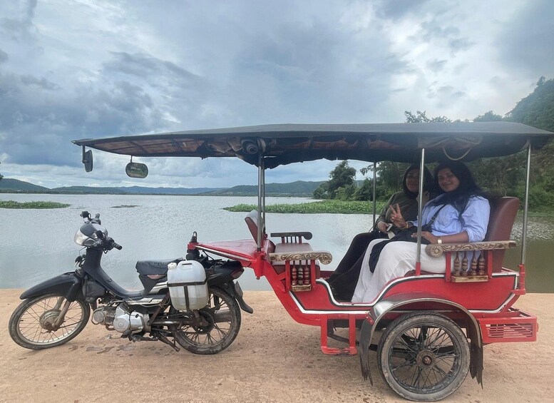 A red rickshaw parked next to a motorcycle by a body of water with mountains in the background. 2