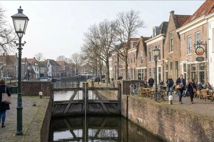 A canal with a wooden bridge, brick buildings, and people walking. 1